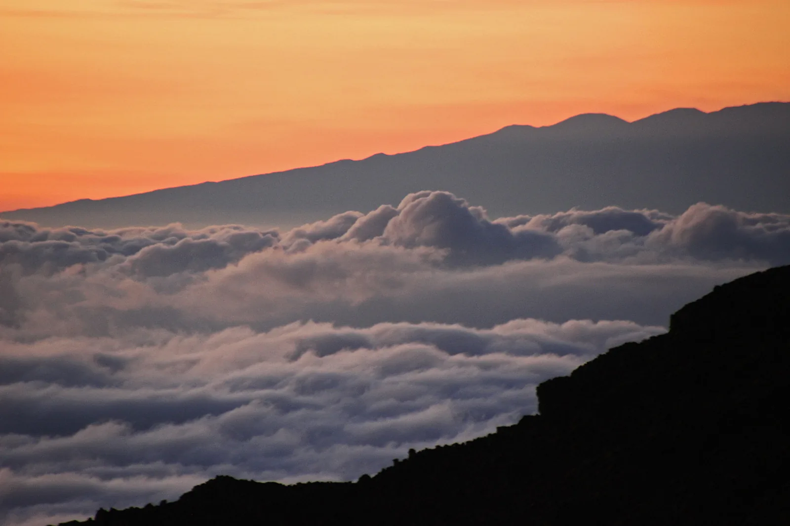 A sea of clouds below the rim of Haleakala at sunrise, with a distant mountain on the horizon