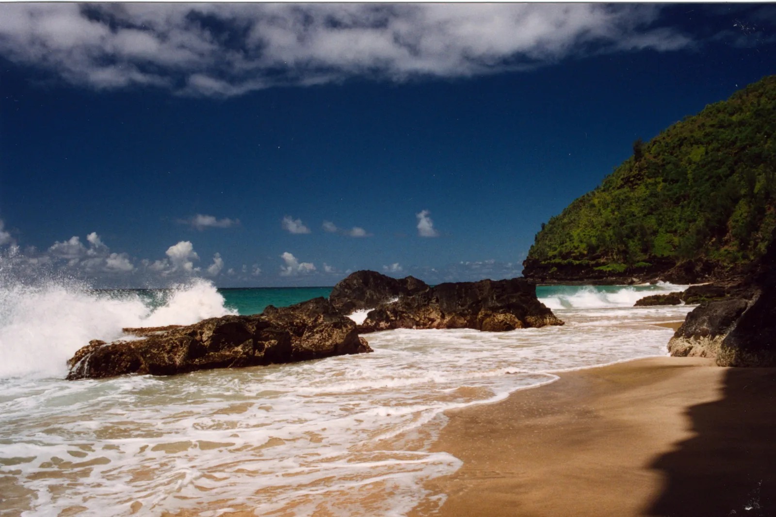 Waves washing over rocks and sand at Hanakapiai Beach below green cliffs on the Kalalau Trail