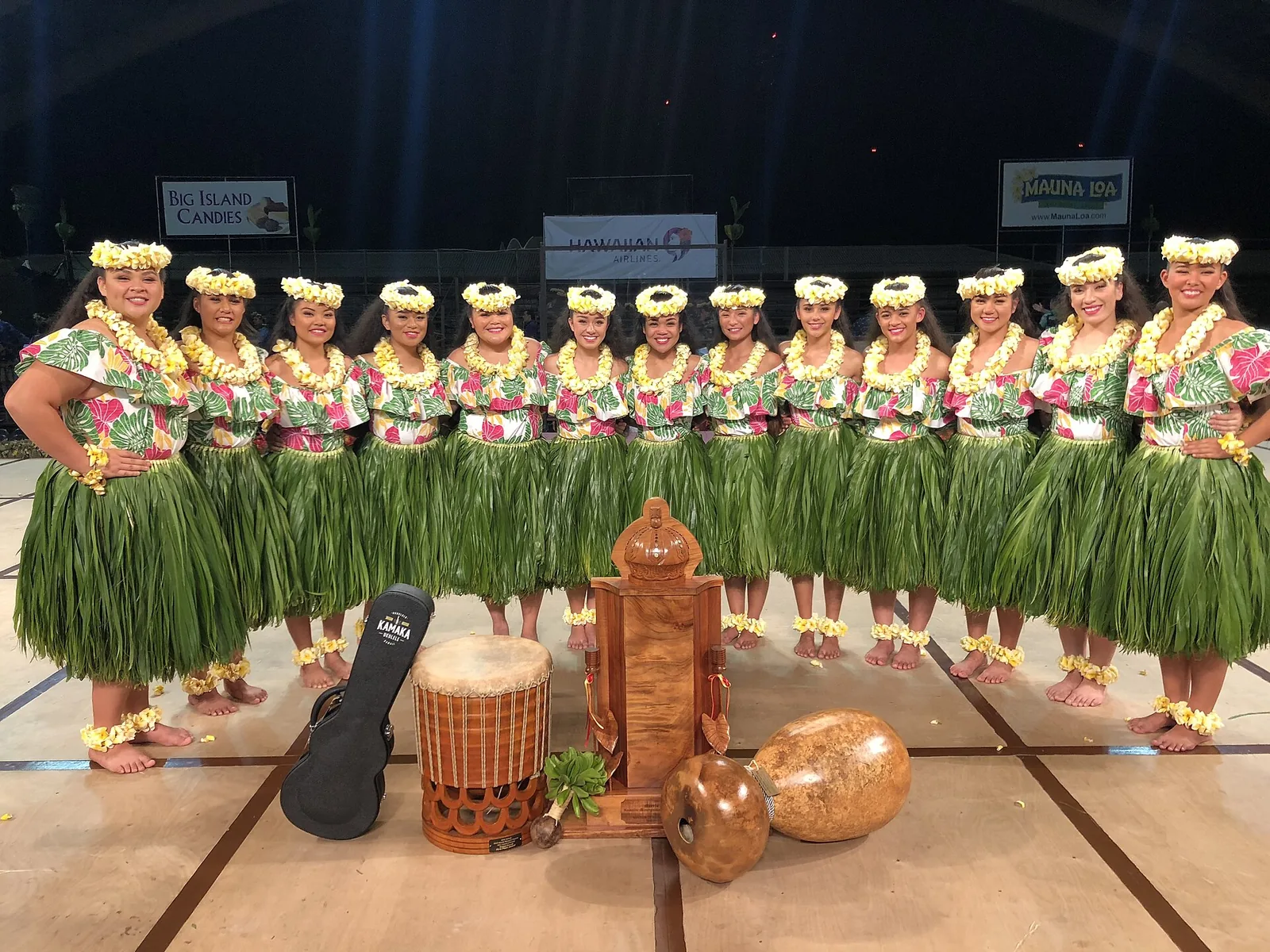A hula halau in ti-leaf skirts and lei standing on stage with drums and instruments at the Merrie Monarch Festival