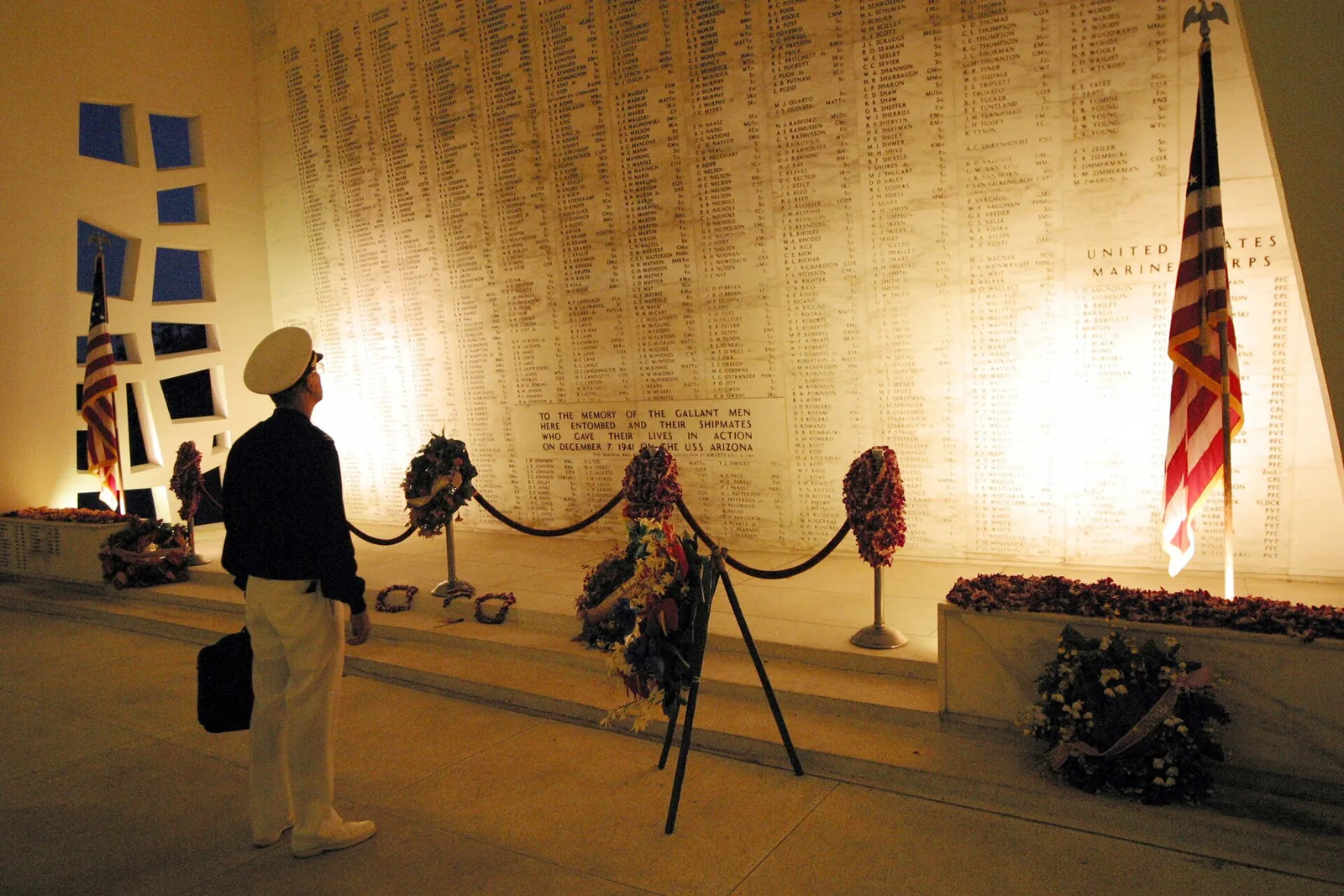 A man in a white cap reading the marble wall of names in the USS Arizona Memorial shrine room, with wreaths and flags