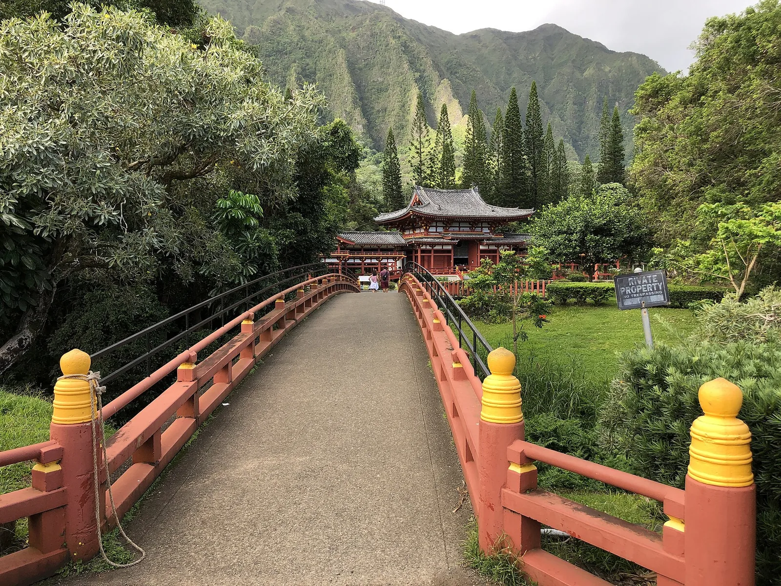 A red-railed footbridge with yellow post caps leading towards the Byodo-In Temple, with green ridges behind