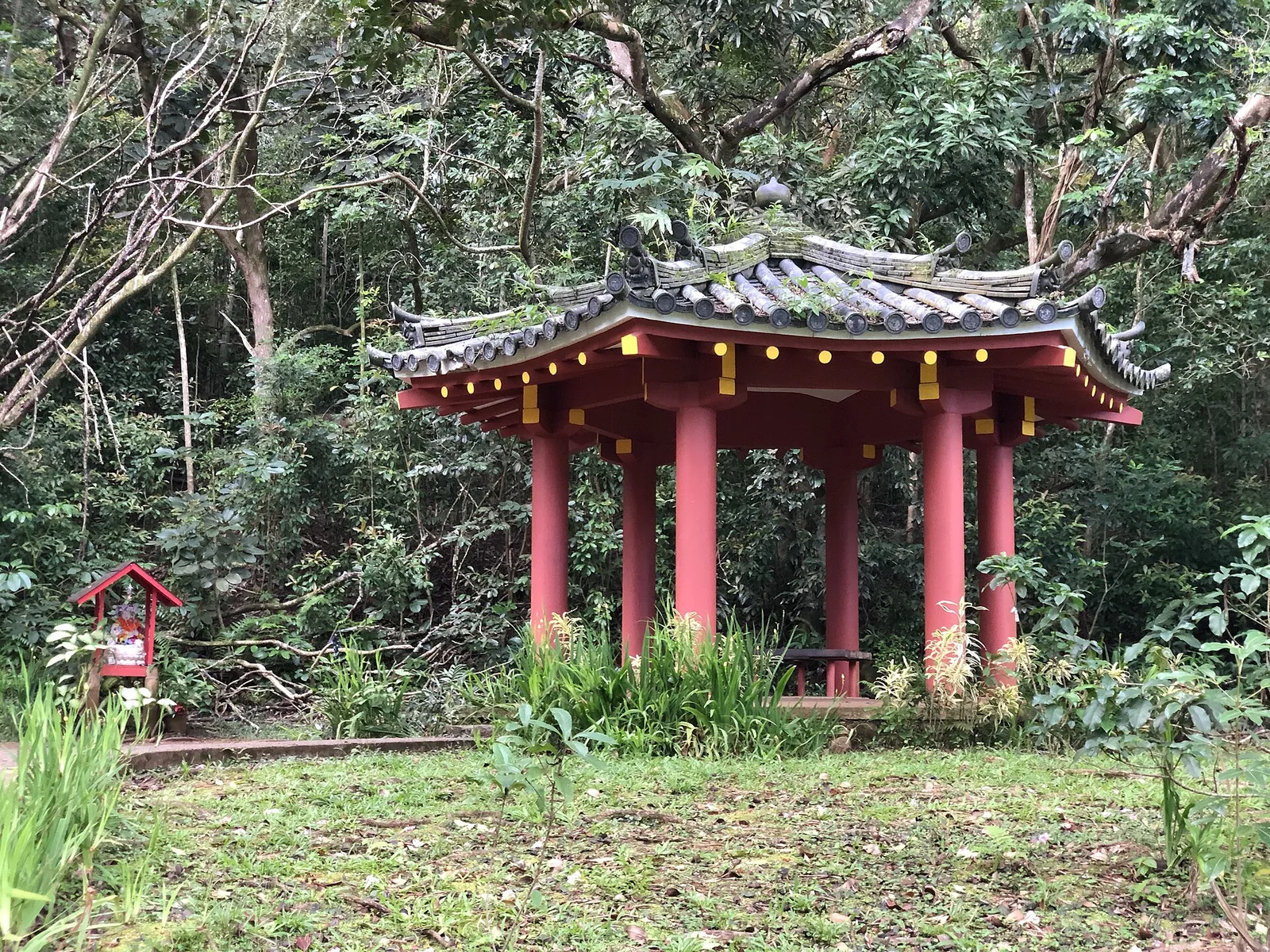 A small red octagonal pavilion with a tiled roof among trees and grass on a slope