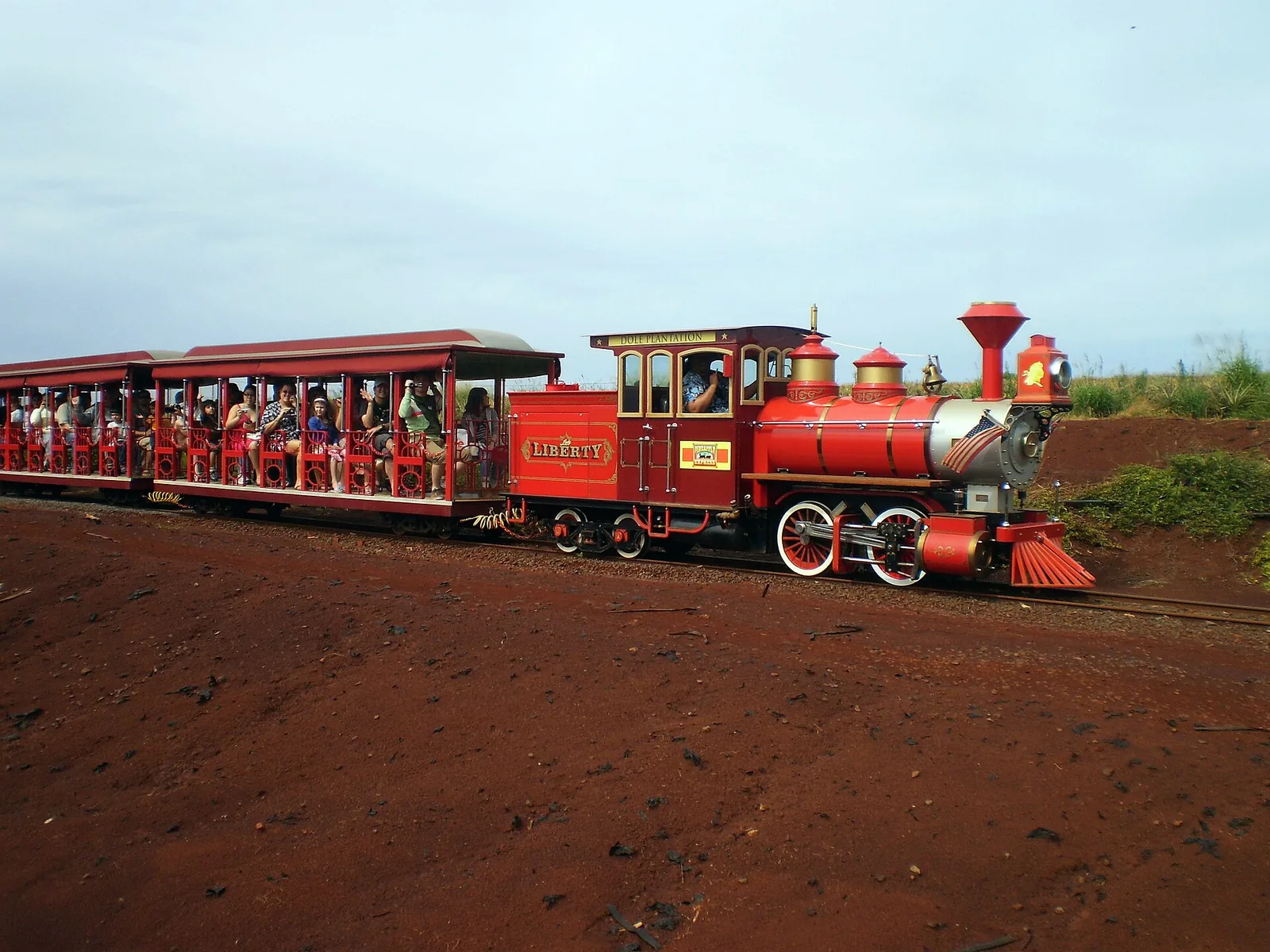 A red steam-style locomotive pulling open carriages full of passengers across red earth at Dole Plantation