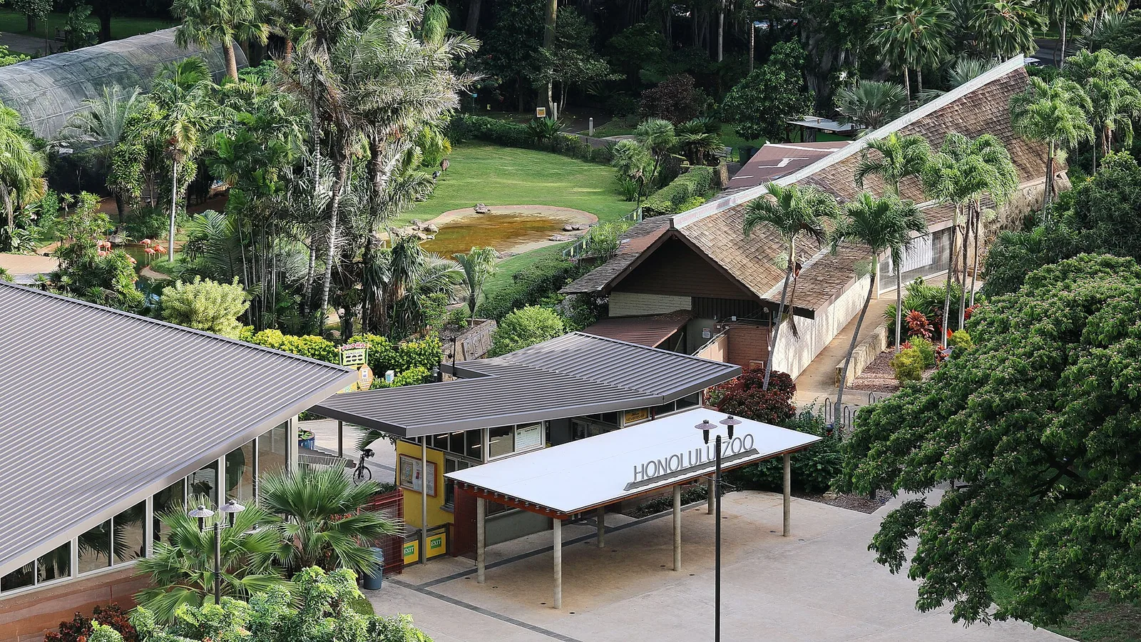 The Honolulu Zoo entrance pavilion and gardens seen from above, with palm trees and exhibit roofs behind