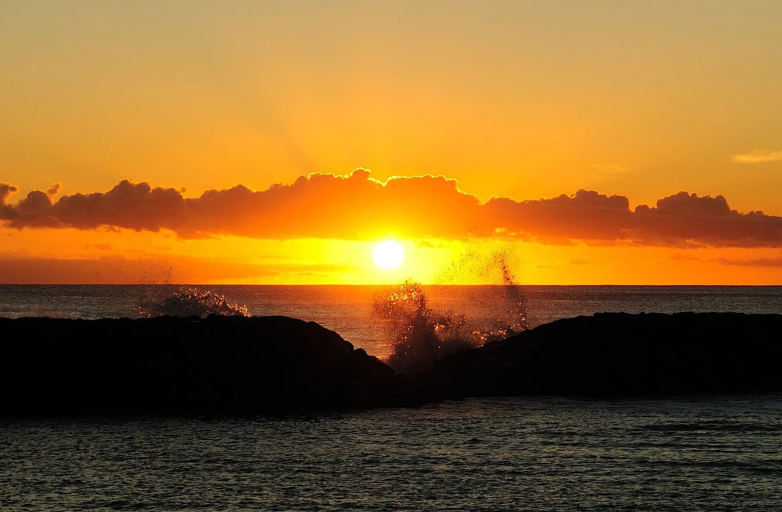 The orange sun setting over the sea behind a dark rock breakwater at Magic Island, with spray in the air