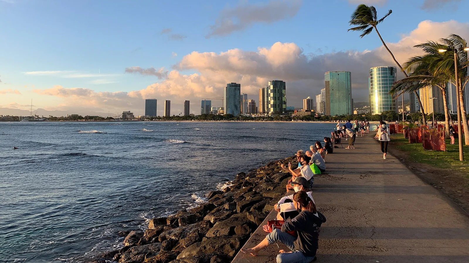 People sitting along the rock wall at Magic Island watching the late sun, with the Kakaako high-rises behind