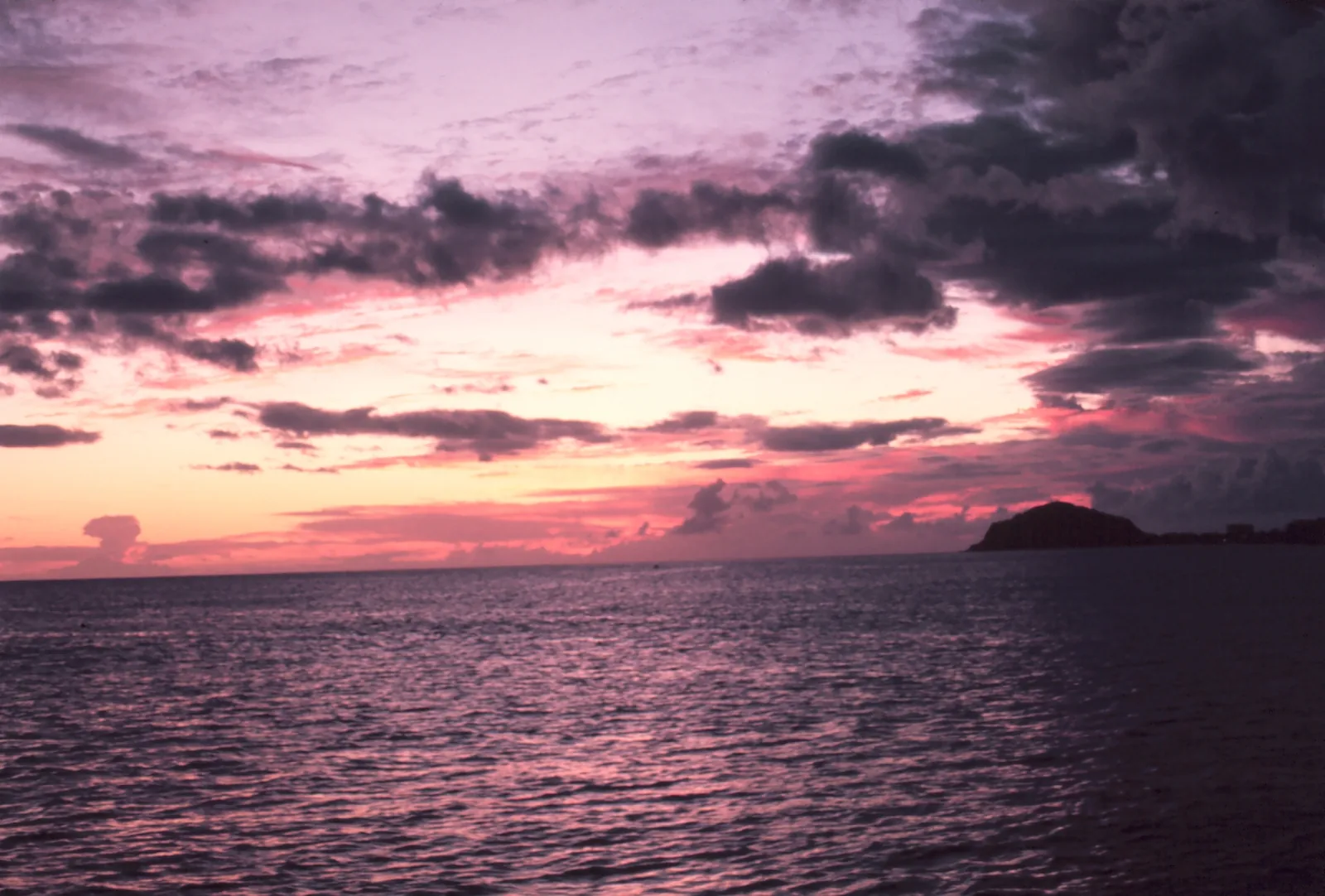 Pink and purple clouds over a dark sea at dusk, with a headland on the horizon off the Waianae coast